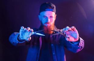 With scissors. Young bearded barber in cap standing in the studio with neon lighting photo