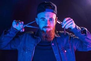 Young bearded barber in cap standing in the studio with neon lighting photo