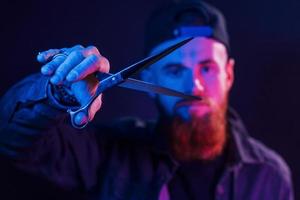 With scissors. Young bearded barber in cap standing in the studio with neon lighting photo