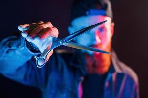 With scissors. Young bearded barber in cap standing in the studio with neon lighting photo