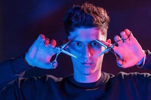 Holds scissors. Young barber with work equipment standing in the studio with neon lighting photo