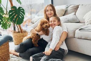 Mother with her daughter playing with dog. Cute little poodle puppy is indoors in the modern domestic room photo