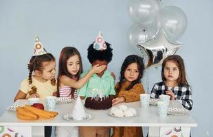 se sienta junto a la mesa. los niños que celebran la fiesta de cumpleaños en el interior se divierten juntos foto