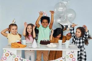 se sienta junto a la mesa. los niños que celebran la fiesta de cumpleaños en el interior se divierten juntos foto