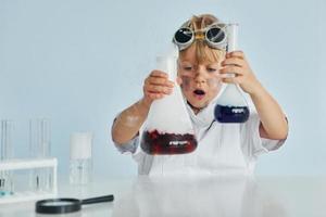 Scared little boy in coat playing a scientist in lab by using equipment photo