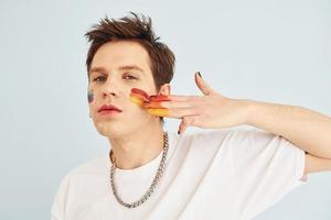 Young gay man is standing in the studio and posing for a camera. Painted by multi colored makeup photo