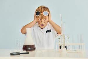 Scared little boy in coat playing a scientist in lab by using equipment photo