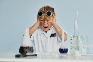 Scared little boy in coat playing a scientist in lab by using equipment photo