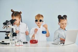 de pie junto a la mesa con tubos de ensayo. los niños con batas blancas juegan a los científicos en el laboratorio usando equipos foto