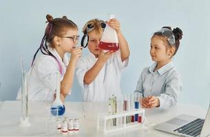 de pie junto a la mesa con tubos de ensayo. los niños con batas blancas juegan a los científicos en el laboratorio usando equipos foto