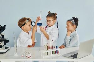 de pie junto a la mesa con tubos de ensayo. los niños con batas blancas juegan a los científicos en el laboratorio usando equipos foto