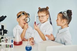 de pie junto a la mesa con tubos de ensayo. los niños con batas blancas juegan a los científicos en el laboratorio usando equipos foto