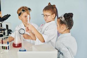 de pie junto a la mesa con tubos de ensayo. los niños con batas blancas juegan a los científicos en el laboratorio usando equipos foto