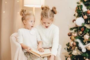 2 blonde girls are sitting on an armchair by the Christmas tree and reading a book. cozy evenings with a book by the tree. The emotion of surprise in children photo