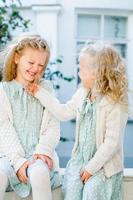 Two little cute girls are sitting in the garden and sharing secrets. One girl whispers something in another's ear. The two sisters have a close relationship. photo