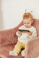 a little girl with ponytails sits on a chair with a phone in her hands. Gadgets from childhood photo