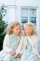 Two little cute girls are sitting in the garden and sharing secrets. One girl whispers something in another's ear. The two sisters have a close relationship. photo