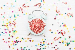 Christmas background with candy canes on alarm clock and multicolored sequins on wooden background photo