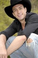 apuesto vaquero se pone en cuclillas para un divertido retrato campestre con un gran sombrero de vaquero negro y una sonrisa. foto