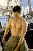 Handsome shirtless muscular captain poses by a sailboat pulling on the ropes with sails drawn. photo