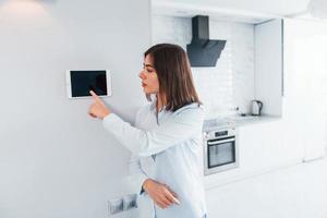 Uses tablet that is mounted in the wall. Young woman is indoors in smart house room at daytime photo