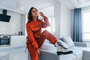 Posing for a camera in red costume. Young woman is indoors in room of modern house at daytime photo