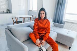 Posing for a camera in red costume. Young woman is indoors in room of modern house at daytime photo