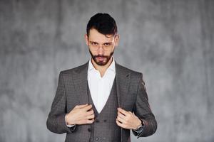 de pie contra el fondo gris. joven y elegante hombre de negocios con traje en el interior. concepción del éxito foto