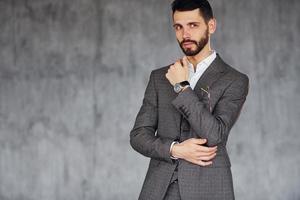 de pie contra el fondo gris. joven y elegante hombre de negocios con traje en el interior. concepción del éxito foto