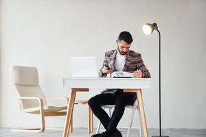 se sienta junto a la mesa en la oficina. joven y elegante hombre de negocios con traje en el interior. concepción del éxito foto