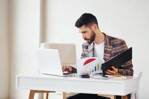 Uses laptop. Young stylish businessman in suit indoors. Conception of success photo