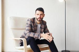 Sits on the chair. Young stylish businessman in suit indoors. Conception of success photo