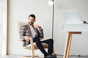 Sits by table in office. Young stylish businessman in suit indoors. Conception of success photo