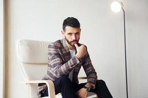 Sits on the chair. Young stylish businessman in suit indoors. Conception of success photo