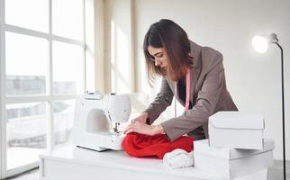 Seamstress works with red cloth. Young woman in formal clothes is indoors. Conception of style photo