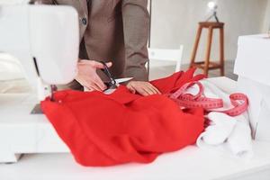 Close up view of seamstress that works with red cloth. Young woman in formal clothes is indoors photo