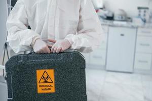 Holds case with biological hazard. Female medical worker in protective uniform works on COVID-19 vaccine photo