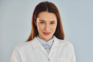 Young female doctor in white coat is standing indoors against blue background photo