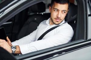 Successful man. Young guy in white shirt is sitting inside of a modern new automobile photo