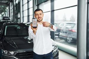 Holds phone with labels and icons. Young man in white shirt is indoors with modern new automobile. Remote control photo