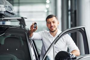 Holds keys. Young man in white shirt is indoors with modern new automobile photo