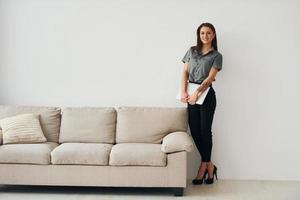 Woman in formal clothes standing near sofa indoors against white wall photo