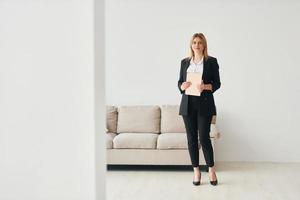 Woman in formal clothes standing near sofa indoors against white wall photo