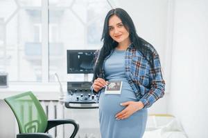 Holds results of ultrasonic scan. Pregnant woman is in the clinic in cabinet and have health check photo