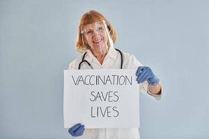 Vaccination saves lives banner. Senior female doctor in white coat is standing indoors photo