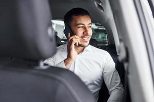 Talks by phone. Young man in white shirt is sitting inside of a modern new automobile photo