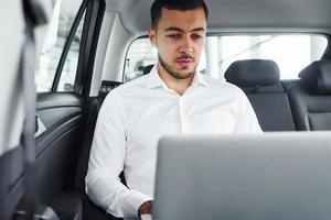 Works by using laptop. Young man in white shirt is sitting inside of a modern new automobile photo