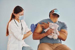 Scared guy with teddy bear. Female doctor giving injection to african american man at hospital photo