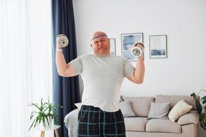 Uses dumbbells. Funny overweight man in casual clothes is indoors at home photo