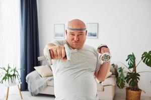 Uses dumbbells. Funny overweight man in casual clothes is indoors at home photo